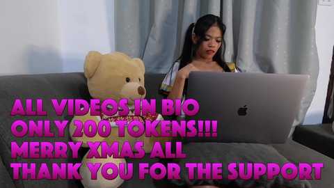 A video shows an Asian woman with long black hair, wearing a red top, working on a laptop in a cozy living room. A large teddy bear sits beside her. Text overlay: \"ALL VIDEO IN BIO ONLY 200 TOOKENS!!! BUY XMAS ALL THANK YOU FOR THE SUPPORT.\