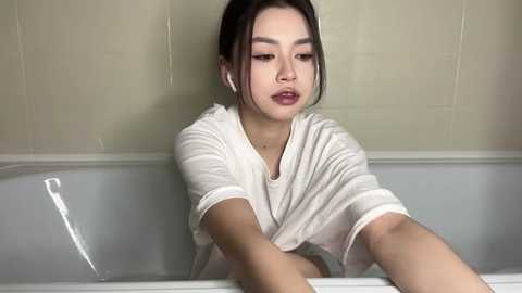 Video of an Asian woman with dark hair in a white t-shirt, leaning against a beige-tiled bathtub, looking contemplative.