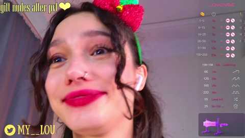 A close-up video of a woman with fair skin, dark curly hair, wearing a green headband with a strawberry, red lipstick, and a nose ring. She has a slight smile.
