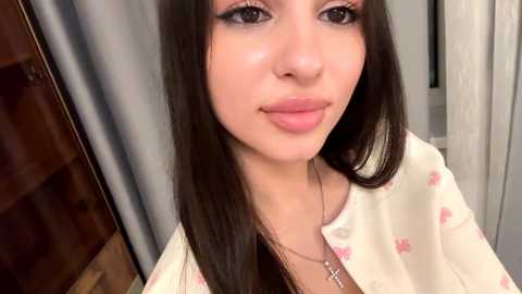A close-up video of a young woman with fair skin, straight dark brown hair, and makeup featuring winged eyeliner. She wears a light-colored, floral-patterned robe with a cross necklace. The background includes wooden furniture and a white curtain.