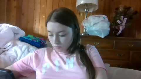 Video of a young girl with straight, dark hair in a ponytail, wearing a light pink t-shirt, sitting on a bed in a wooden-paneled room with a white lamp, colorful clothes, and a stuffed animal.