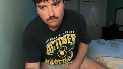 Video of a young man with a mustache wearing a black \"Illinois State University\" t-shirt, sitting on a bed in a dimly lit room with teal walls and white doors.
