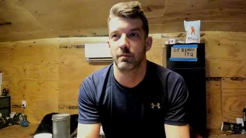 Video of a muscular white man with short brown hair and a beard, wearing a black Under Armour shirt, sitting in a wooden cabin. Background includes a white AC unit, a sign reading \"Of Kiny 1/2,\" and various wooden objects.