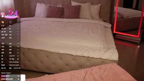 Video of a modern bedroom with a large, tufted beige headboard bed, white bedding, and a pink knitted blanket. A nightstand with a pink lamp and a pink toy is visible on the left.