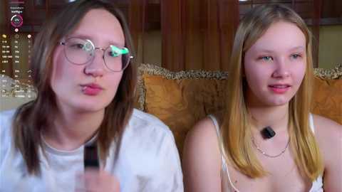 Video of two young, Caucasian girls with glasses, wearing white tops, sitting on a couch with brown cushions.