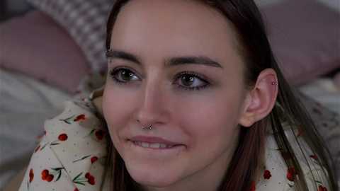Video of a young woman with fair skin, straight brown hair, and a septum piercing, smiling, wearing a floral-patterned shirt, lying on a bed with checkered pillows.