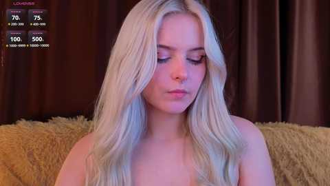 A video of a young, fair-skinned woman with long, platinum blonde hair, wearing minimal makeup, sitting on a yellow plush couch. The background features dark brown curtains and a digital clock display showing 7:25 AM.