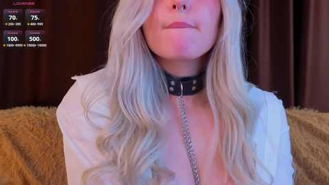 Video of a woman with long, wavy, platinum blonde hair, wearing a black leather collar with a metal chain leash, and a nose ring. She's indoors, in a dimly lit room with brown curtains and a beige fur blanket.