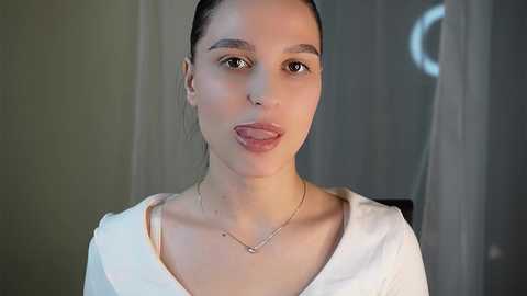 Media: Video of a light-skinned woman with dark hair, wearing a white top and silver necklace, sticking out her tongue, against a blurred gray background.