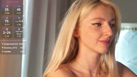 Video of a blonde Caucasian woman with straight hair and a slim physique, wearing a strapless top, standing indoors with a blurred background, displaying a weather app with temperature details.