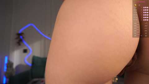 Video of a nude woman's buttocks and lower back, highlighting smooth skin and a hint of a tattoo. The background is dimly lit with blue and purple neon lights, creating a sensual atmosphere.