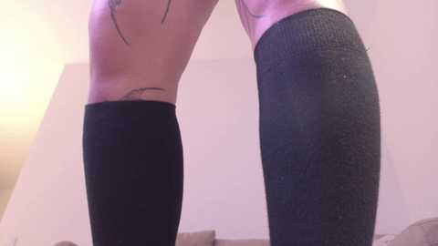 Video of a person's legs in black knee-high socks, standing on a bed with beige sheets, against a pale wall background.