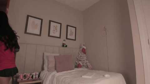 Video of a cozy, minimalistic bedroom with beige walls, a white tufted headboard, three framed botanical prints, a small Christmas tree, and a pink and white bed.