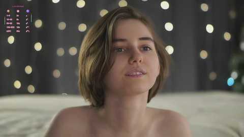 Video of a young, fair-skinned woman with short, light brown hair, gazing directly at the camera. She has a slender build and is topless, set against a blurred, bokeh background of warm, golden lights and dark curtains.