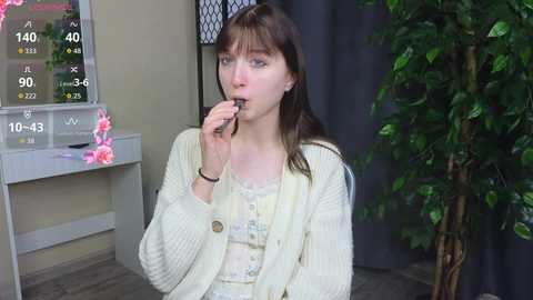 Video of a pale-skinned young woman with dark hair, wearing a cream cardigan, eating a chocolate bar. Background features a white vanity with a floral arrangement, a green potted plant, and a window with a black curtain.