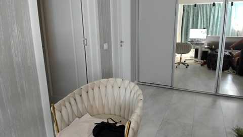 Media: Video of a modern, minimalist room with a cream-colored, tufted armchair, a sliding door mirror, a desk with a monitor, and light grey flooring.