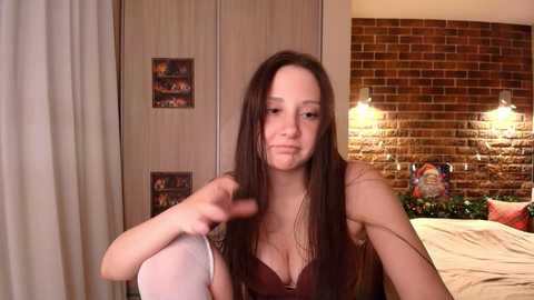 Video of a young woman with long, straight brown hair, wearing a black bra and white stockings, sitting in a bedroom with brick wall and wooden wardrobe.