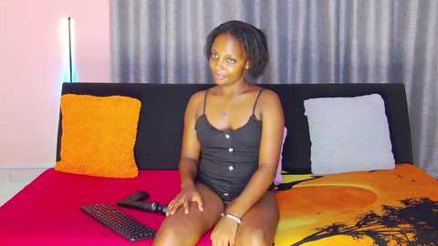 Video of a Black woman with short, curly hair wearing a black camisole, sitting on a bed with orange and yellow pillows. She has a neutral expression.