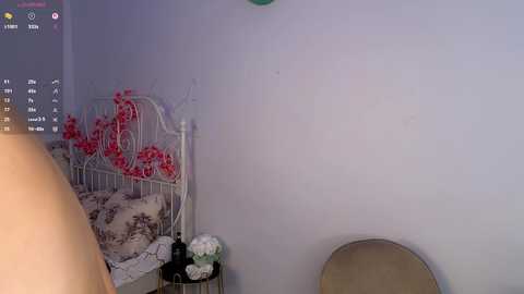 Video of a dimly lit bedroom with a white metal crib adorned with red and white floral sheets, a beige chair, and a white wall.
