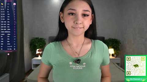 Video of a young Asian woman with straight black hair, wearing a green t-shirt, in a modern, dimly lit room with a TV showing a cryptocurrency exchange screen.