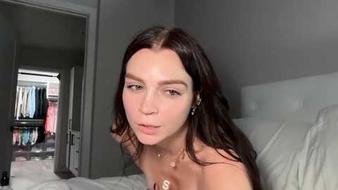 Media: Video of a young woman with long, dark brown hair, fair skin, and light makeup, wearing a gold necklace, lying on a bed in a minimalist, gray-toned bedroom.