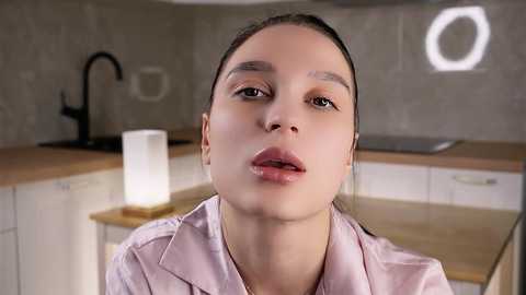 Video of a young woman with light skin, straight brown hair, and full lips, wearing a light pink blouse in a modern, minimalist kitchen with wooden countertops, a black faucet, and a hexagonal light fixture.