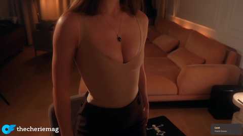 Video of a woman with light skin, wearing a low-cut beige top and black pants, standing in a dimly lit room with a beige sofa and wooden floor.