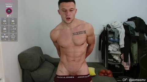 Video of a muscular, light-skinned man in maroon briefs, wearing a tattoo on his right shoulder. Background features a gray couch, messy clothes rack, and a fitness tracker.