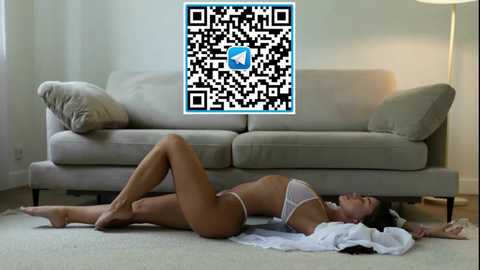 Video of a woman in white lingerie lying on a carpeted floor in front of a beige couch. QR code overlay.