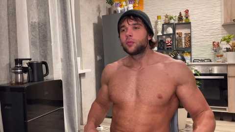 Video of a shirtless, muscular man with a dark green beanie, making a kissy face, in a modern kitchen with stainless steel appliances, white brick wall, and colorful decor.
