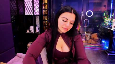 Video of a dark-haired woman in a low-cut burgundy top, sitting in a dimly-lit room with purple accents and a large window displaying holiday decorations.