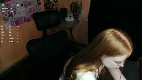 Video of a pale, red-haired woman in a white shirt sitting in a dark room with a black chair, a wall-mounted calendar, and a plant.