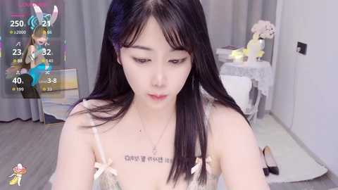 Video of an Asian woman with long black hair and pale skin, wearing a lacy white bra, sitting on the floor in a room with light-colored furniture.