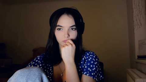 Video of a young woman with long black hair, wearing a blue polka-dot dress, sitting thoughtfully with a hand on her chin, in a dimly lit room with beige walls.
