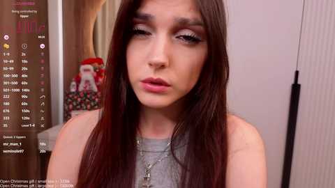 A video of a young woman with long, straight, dark brown hair, wearing a gray top, makeup, and a necklace. Background shows a room with a red, white, and green Santa Claus ornament on a table.