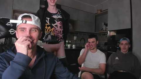 Video of four young men in a dimly lit, modern kitchen. One man, shirtless and wearing a backwards cap, sits on a stool. Others wear casual clothing; one has a tattoo.