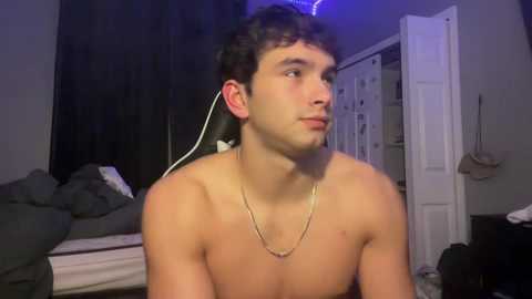 Video of a shirtless young man with curly dark hair, wearing a gold necklace, listening to music with earphones in a dimly lit bedroom with a bed, white door, and cluttered dresser.