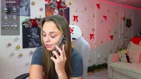 Video of a young woman with long brown hair, wearing a blue shirt, talking on a smartphone in a festive, decorated room with holiday ornaments and a Santa hat.