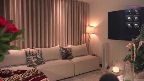 Video of a cozy living room with beige sofa adorned with festive pillows, beige curtains, warm lighting, Christmas tree, and a large TV screen displaying a score board.