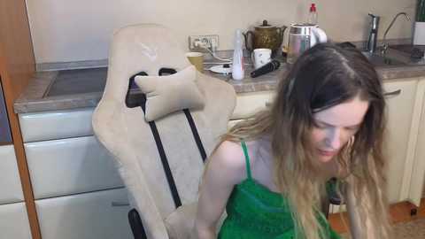 Video of a young woman with wavy, light brown hair in a green dress, seated on a beige gaming chair in a kitchen, surrounded by cooking utensils and appliances.