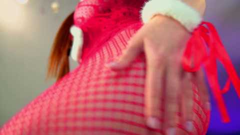 A close-up video of a woman's lower torso in a red fishnet bodysuit with white fluffy trim. Her hand rests on her hip, fingers spread, revealing a glimpse of her fair skin and red fingernails.