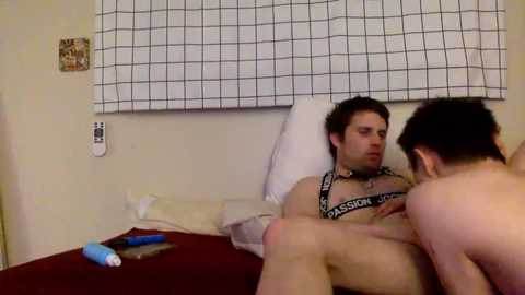 Video of a naked man with a tattooed chest lying on a bed with a white pillow, a woman performing oral sex on him, a blue vibrator, and a TV on the beige wall.