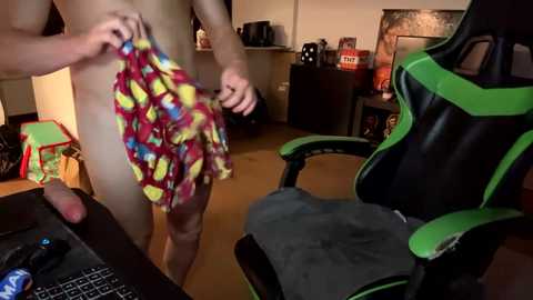 A video of a naked man with fair skin wearing colorful, cartoon-patterned boxers, standing in a dimly lit room with a green and black gaming chair and a cluttered desk.
