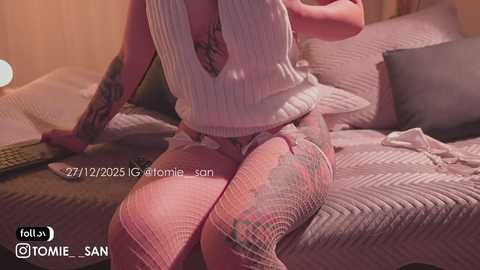 Video of a tattooed woman in a white ribbed top and fishnet stockings, seated on a bed with gray bedding, holding a phone.