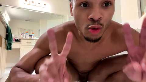 Media: Video of a shirtless, light-skinned man with a trimmed beard and short hair, making a peace sign with both hands, in a bathroom with white cabinets and a mirror.