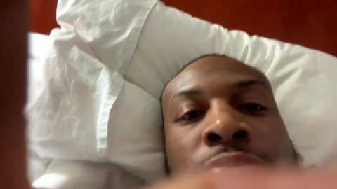 Video of a Black man lying on a white pillow, eyes closed, with a reddish-brown background, suggesting a warm, cozy setting.