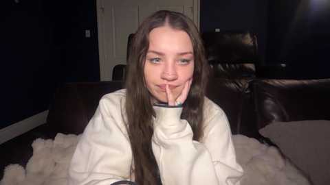 Video of a young Caucasian woman with long brown hair, wearing a white hoodie, sitting on a white furry rug in a dimly lit room with dark walls and leather furniture.