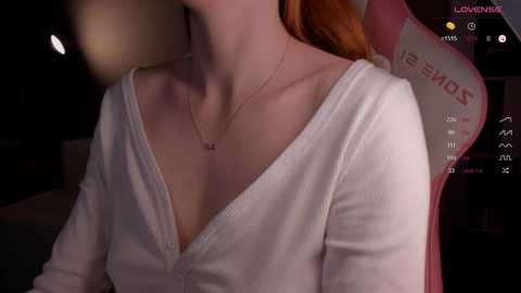 A dimly-lit video of a pale-skinned woman with long red hair, wearing a low-cut white cardigan and a delicate necklace, seated in a dark room with a pink gaming chair.
