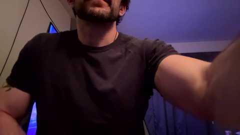A video of a muscular, light-skinned man with a trimmed beard and mustache, wearing a dark T-shirt, taking a selfie indoors. The background shows a dimly lit room with white walls and a blue-tinted window.