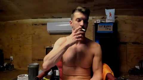 Video of a shirtless, muscular, light-skinned man with short, styled hair, smoking a cigarette, seated in a cluttered, dimly lit wooden-paneled room with a mounted air conditioner and a sign reading \"OFF DUTY 1972.\